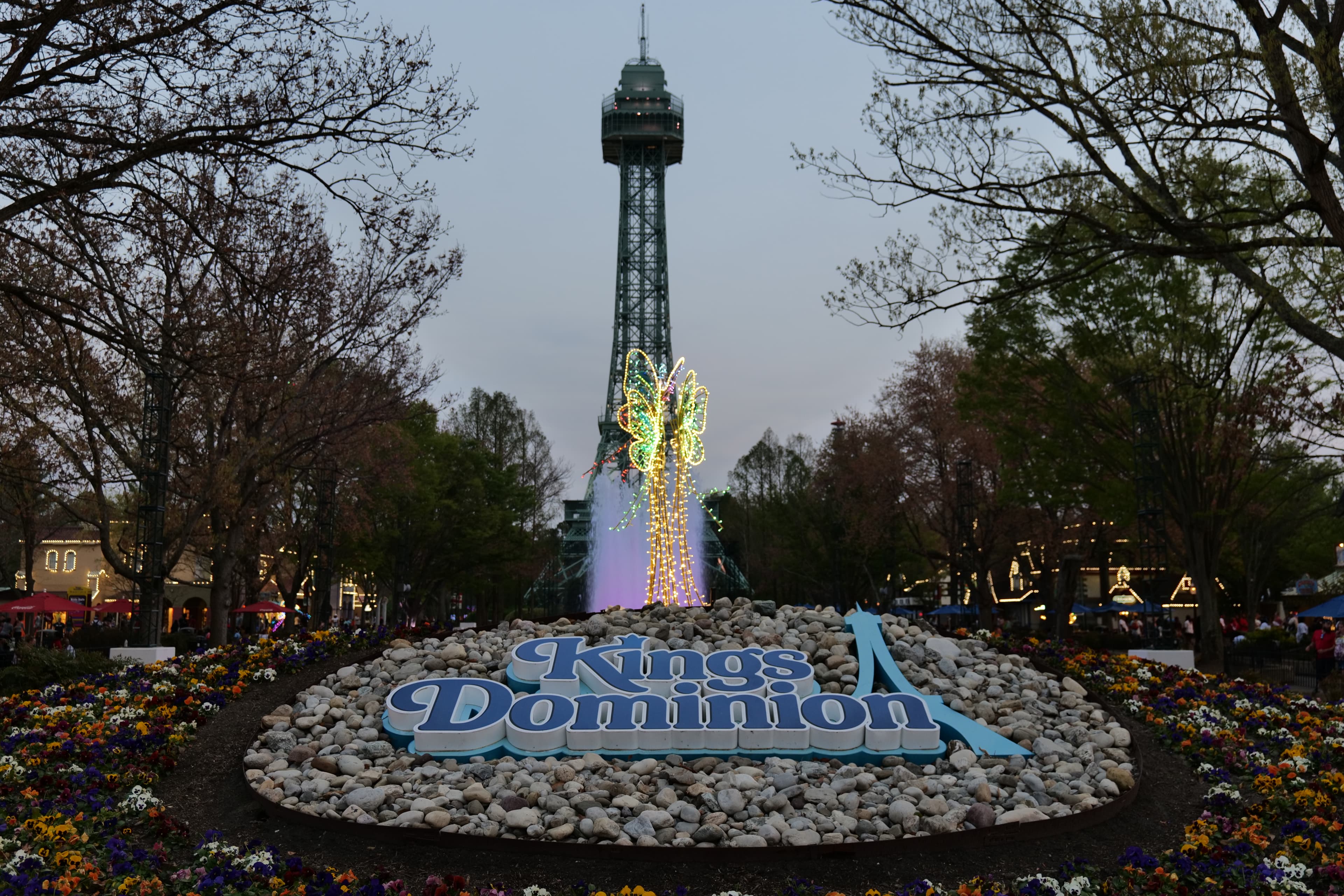 Kings Dominion sign among flowers, with a fountain and lattice tower behind it.